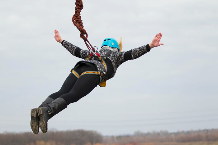 Person wearing blue helmet and harness bungee jumping outdoors with arms outstretched against overcast sky.