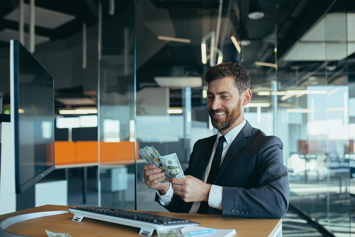 A male lawyer in a suit counting cash with a smile inside a modern office, reflecting unexpected legal insights.