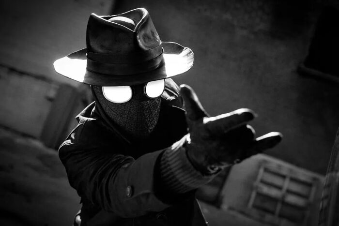 Spider-Noir in dark trench coat and hat, with glowing eyes, posed in moody black and white setting, showcasing powers.
