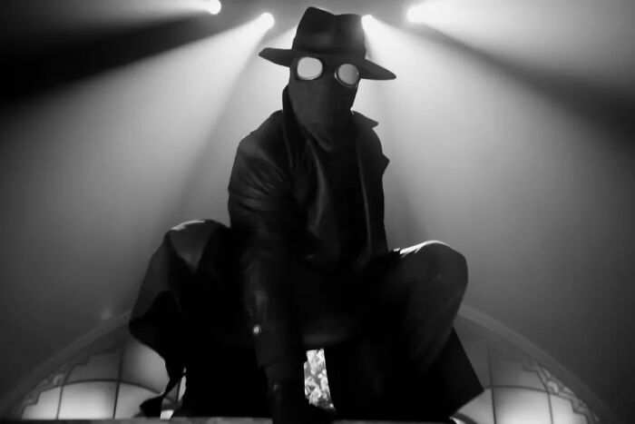 Spider-Noir crouching in a dark, smoky room wearing a trench coat, fedora, and round reflective goggles.
