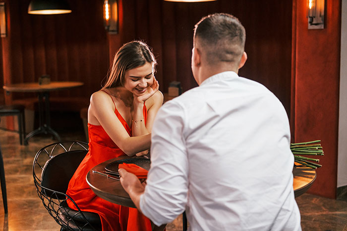 Man plans proposal at restaurant while woman in red dress smiles, unaware of the ring ending up in another woman's dessert.