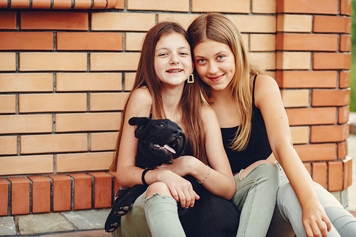 Two teenage girls sitting by a brick wall with a black dog, showing a close stepdaughters relationship despite cheating and lies.