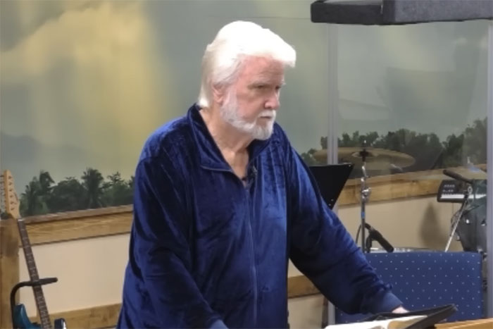 Older man with white hair and beard wearing a blue jacket, standing by music instruments indoors in a casual setting.