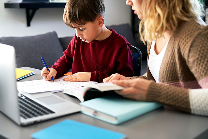 Young boy focused on writing while adult woman helps with homework, illustrating learning and conspiracy theories concept.