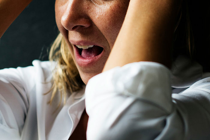 Close-up of a shocked woman in a white shirt reacting with hands on her head about conspiracy theories and disbelief.