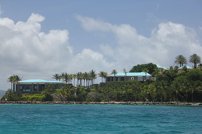 Coastal view of luxury beachfront homes surrounded by palm trees under a cloudy blue sky, ocean in foreground.