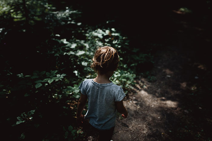 Young child walking alone on a shadowed forest path surrounded by dense greenery, evoking mystery and curiosity.