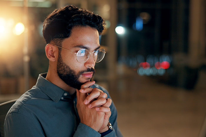 Man with glasses and beard concentrating deeply while pondering conspiracy theories late at night in an office setting