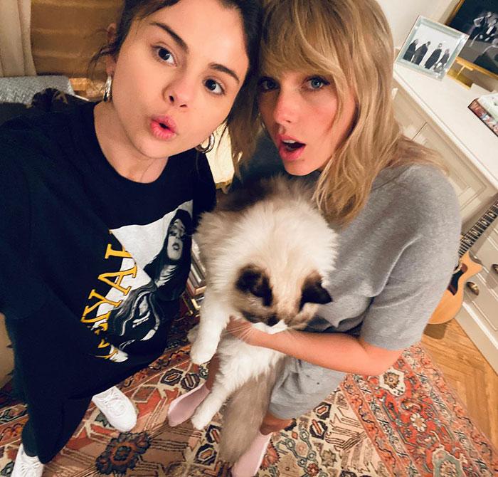Selena Gomez and Taylor Swift posing indoors with a cat, capturing a moment amid gift backlash rumors.