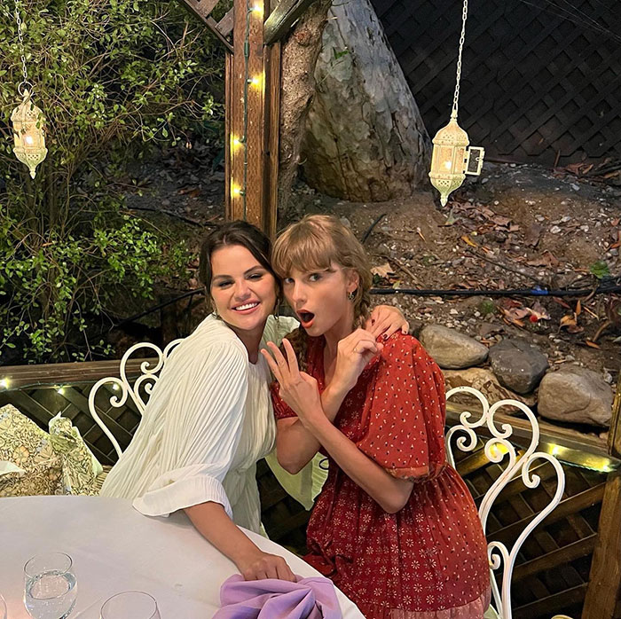 Selena Gomez and Taylor Swift posing together outdoors, lighting decorations visible, capturing a candid moment.