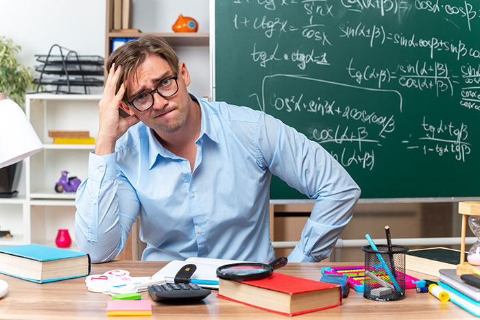 Stressed teacher sitting at desk in classroom with math formulas on chalkboard, dealing with blacklisting and coworker threats.