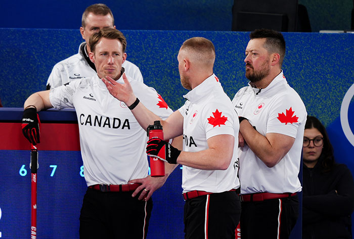 Canadian curling team members in white uniforms with red maple leaf discussing strategy during Olympics event.