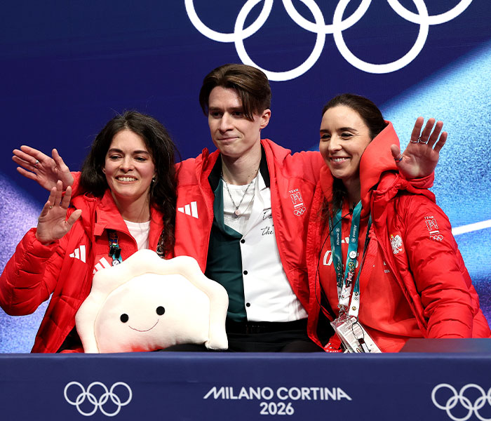Three Winter Olympics athletes in red jackets posing with a smiling dumpling plush at Milano Cortina 2026 event.