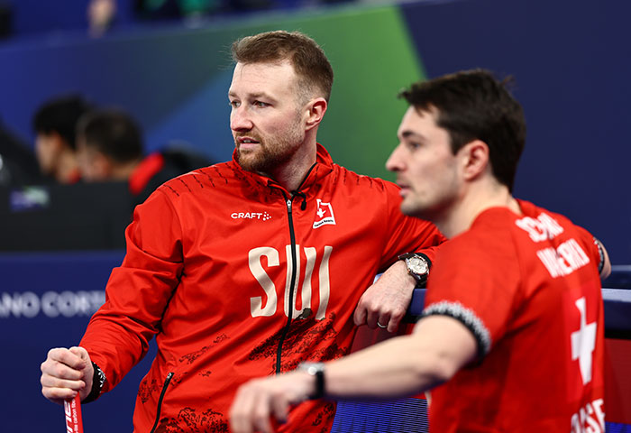 Two male athletes in red Swiss team jackets sitting at the Olympics after a major change following cheating accusations.