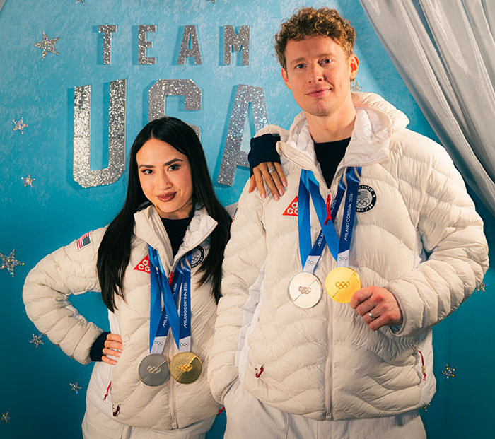 Two Olympic ice skaters wearing white jackets and medals posing in front of a Team USA backdrop amid French ice skaters scandal