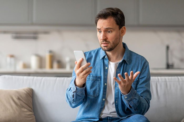 Man in casual clothes sitting on sofa looking shocked at phone, reflecting troubled marriage and therapy issues.