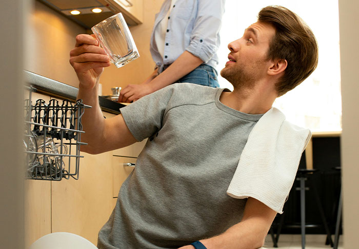 Man-child behavior example, young man loading dishwasher casually with towel over shoulder in kitchen setting.