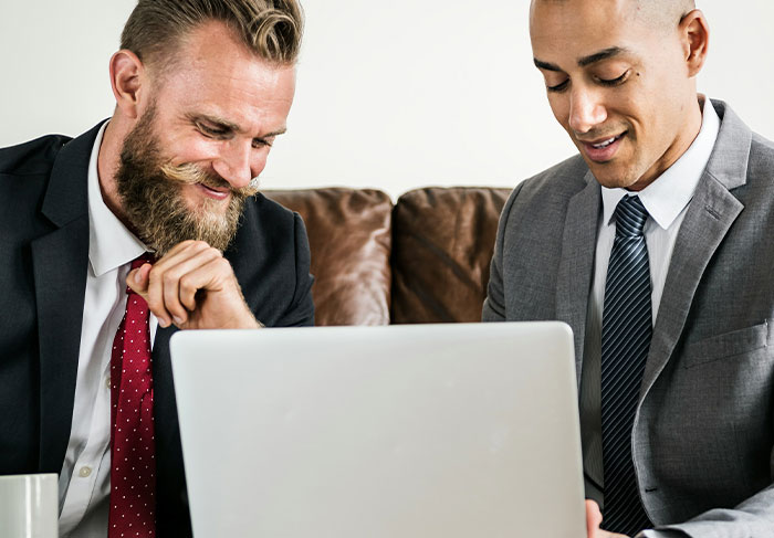 Two businessmen in suits smiling while looking at a laptop, discussing man-child behaviors in a professional setting.