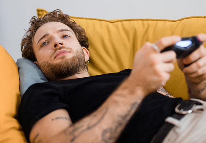 Young man with tattoos and nose ring lying on a couch playing video games, showing man-child behavior traits.