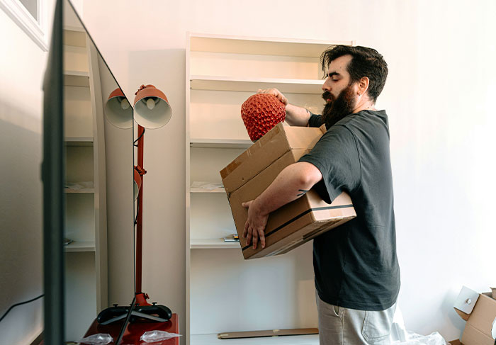 Man carrying boxes and a lamp while unpacking, illustrating behaviors of a man-child in a home setting.