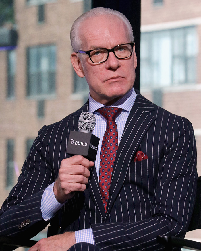 Tim Gunn speaking at an event wearing glasses and a striped suit, sharing about celibacy and personal experiences.