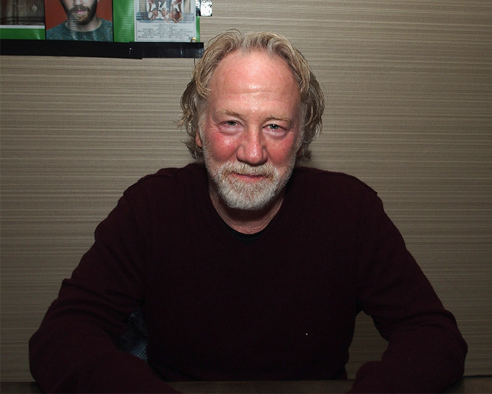 Timothy Busfield with gray hair and beard, sitting indoors wearing a dark sweater in a casual setting.
