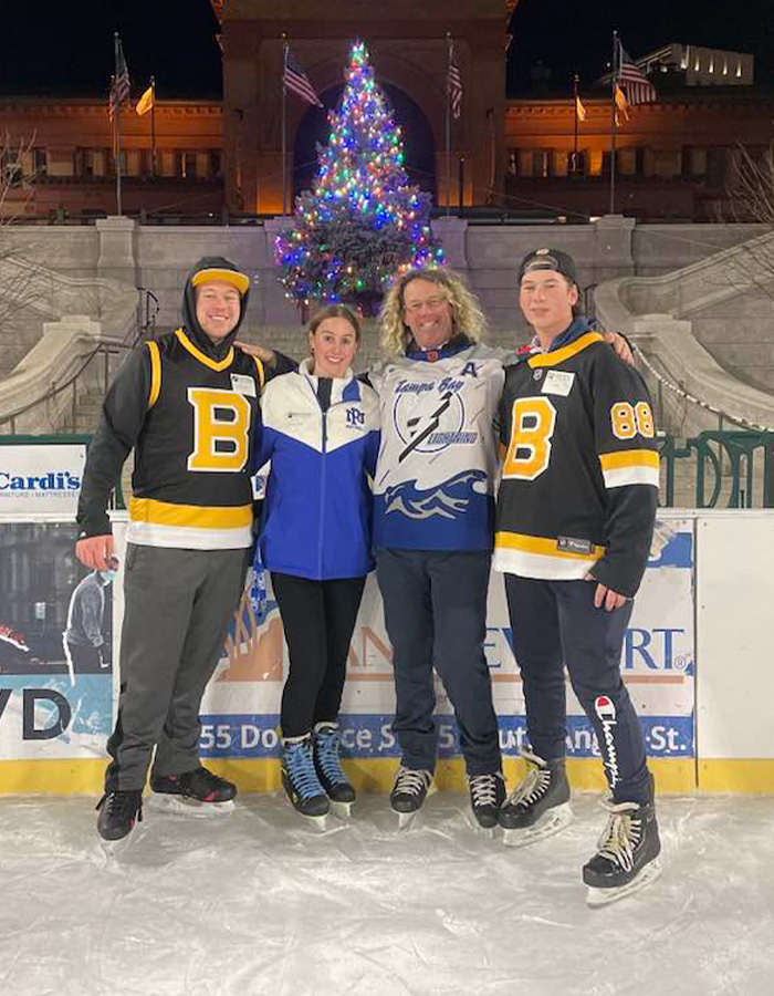 Group of four people ice skating at night with a decorated Christmas tree in the background at an ice rink setting.