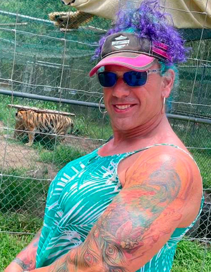 Person with colorful hair and tattoos smiling outdoors near fenced area with a tiger, related to ice rink attacker family discovery.