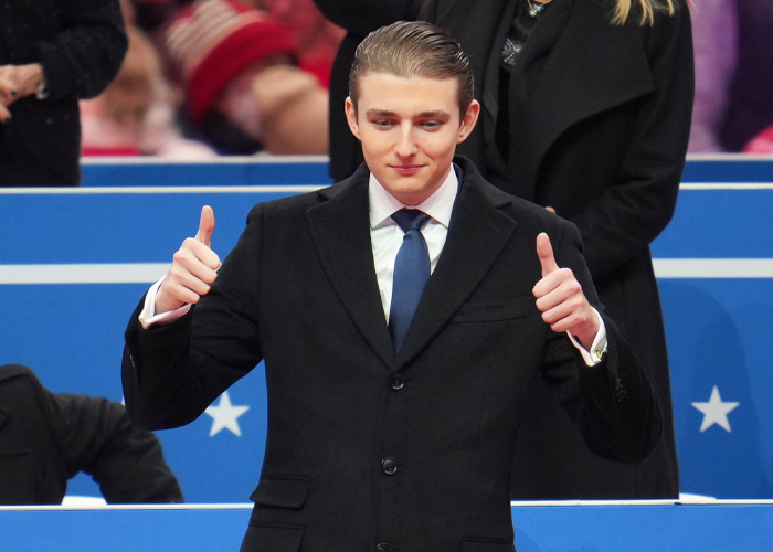 Barron Trump in a black suit and blue tie giving double thumbs up gesture at a public event with spectators behind him.