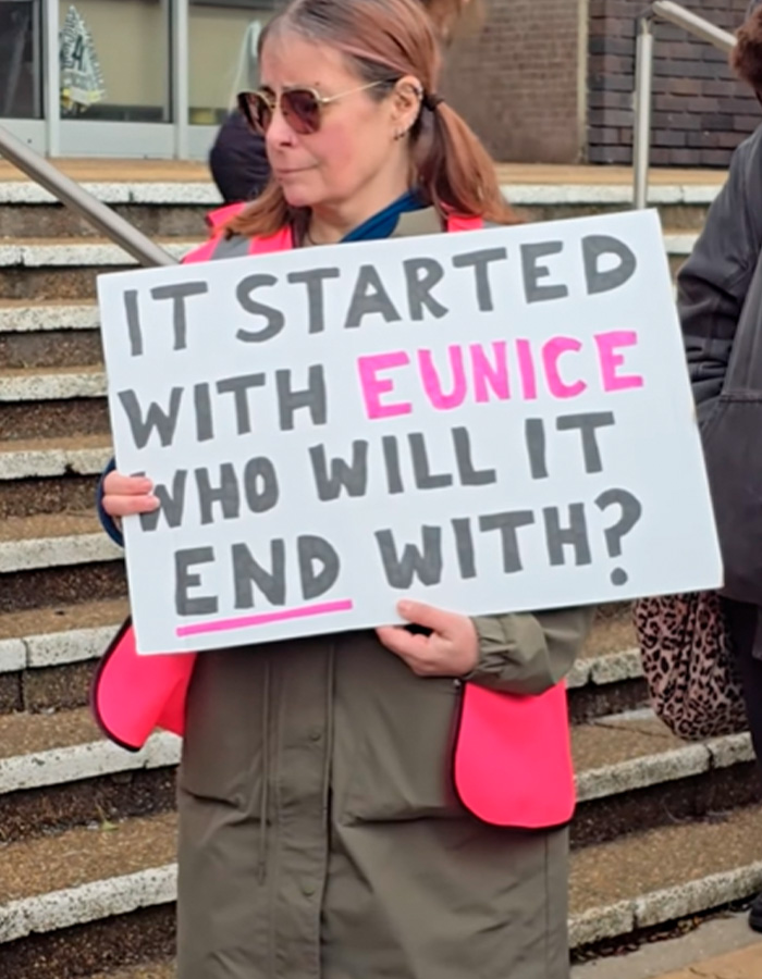 Woman wearing sunglasses and a pink vest holding a sign about two students involved in fireworks sheep incident karma.
