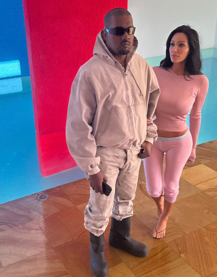 Bianca Censori com blusa e calça estilo lingerie rosa ao lado de um homem vestido com roupa casual bege dentro de casa.