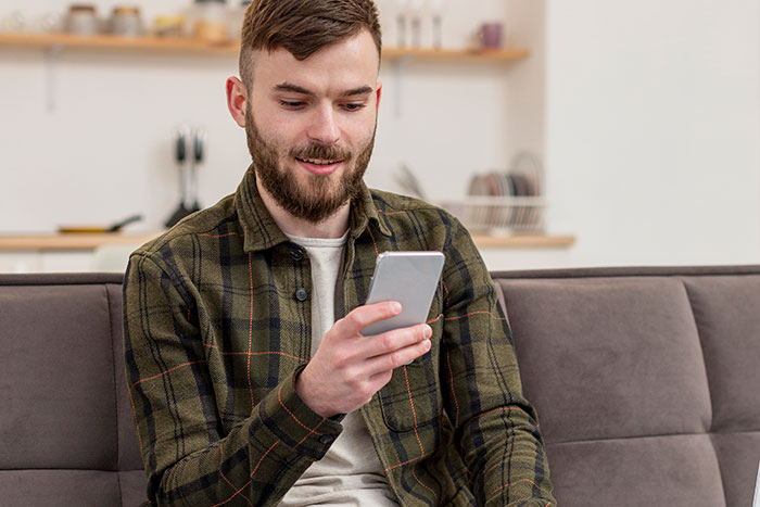 Young man sitting on a couch, smiling and looking at his smartphone, illustrating responding to weaponized incompetence.