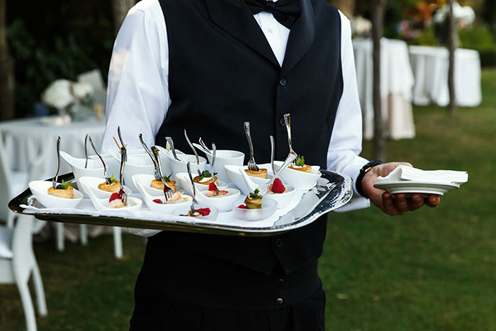 Waiter holding tray of appetizers at outdoor wedding catering event with elegant white table settings in background Waiter holding tray of appetizers at outdoor wedding catering event with elegant white table settings in background