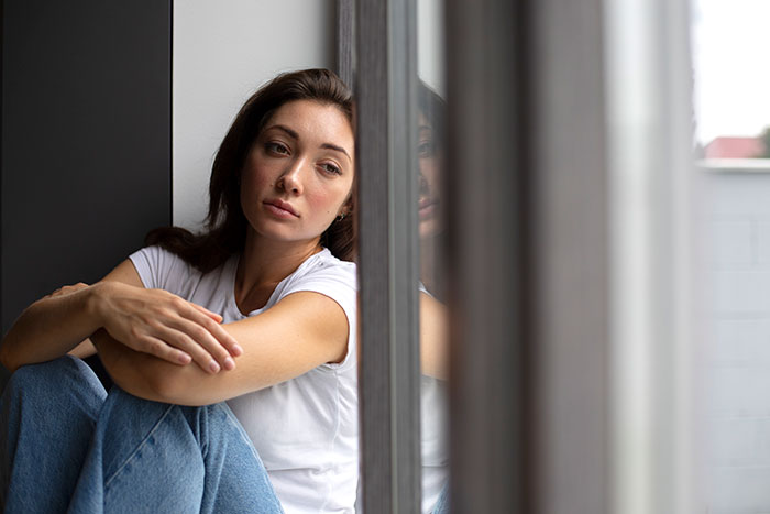 A thoughtful woman in a white shirt and jeans sitting by a window reflecting on reasons people give to have kids.
