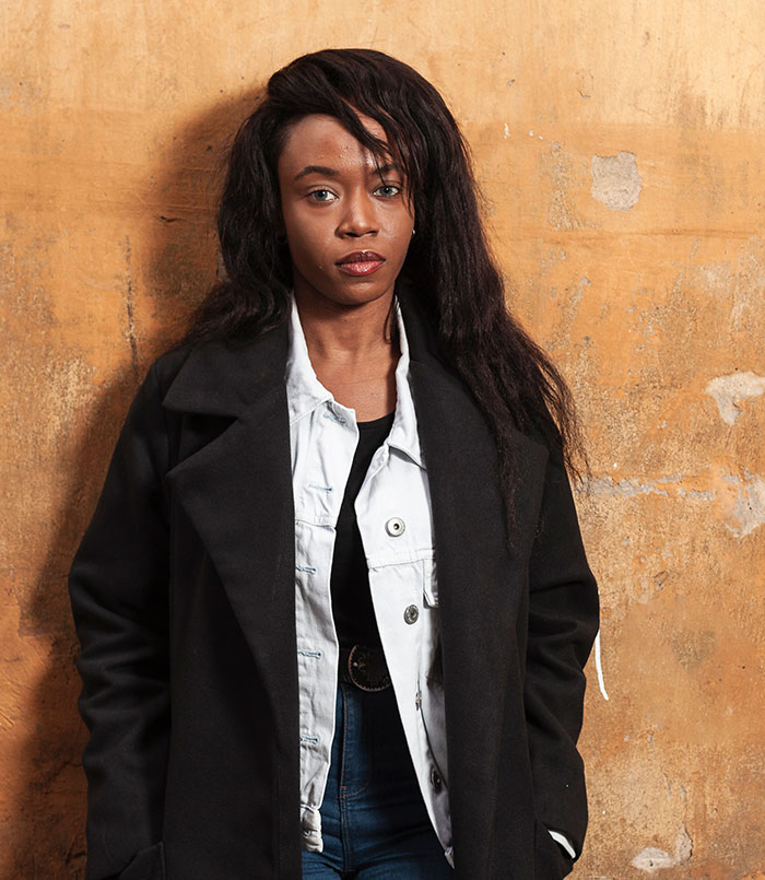 Young woman in layered black and white jacket standing against a textured wall, reflecting on reasons people have kids.