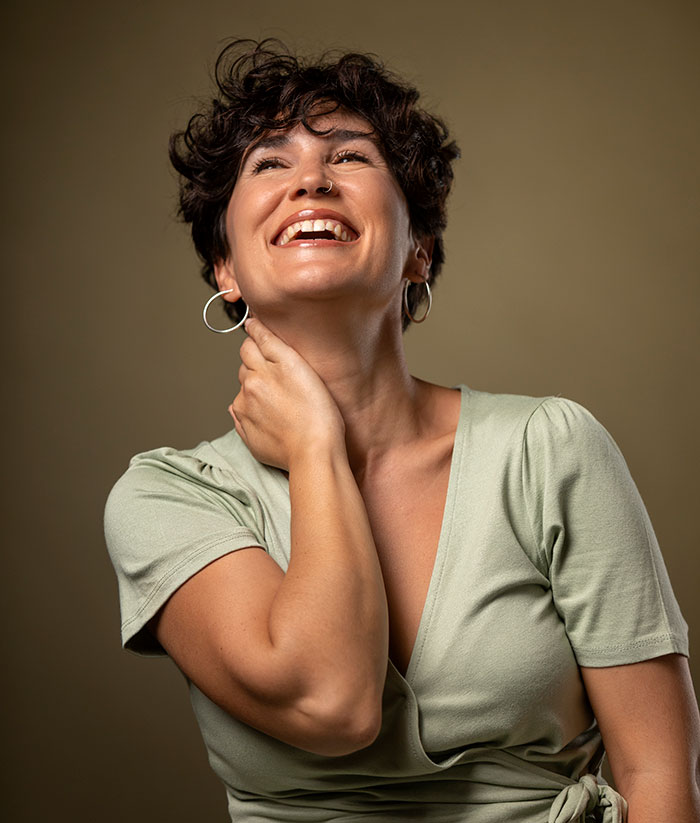 Smiling woman with short curly hair and hoop earrings, looking up while touching her neck in a casual green top.