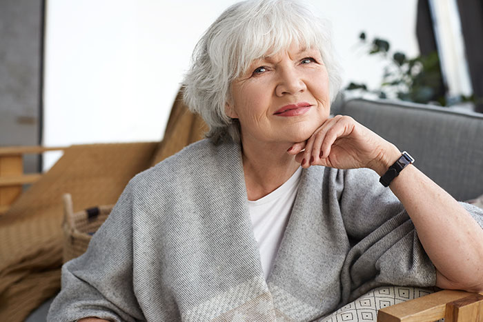 Older woman with white hair sitting on a couch, wrapped in a gray shawl, reflecting on reasons to have kids.