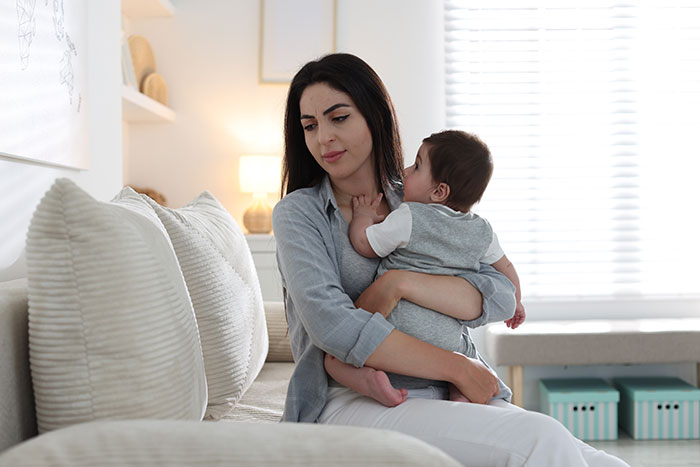 Woman holding baby on couch, reflecting on reasons people give to have kids in a bright, cozy living room.