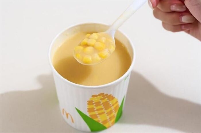 Close-up of a hand holding a spoon with corn soup from a McDonald's cup, showcasing unique food items in different countries.