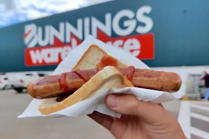 Hand holding a sausage in bread with sauce outside a warehouse store, showcasing common things across countries.