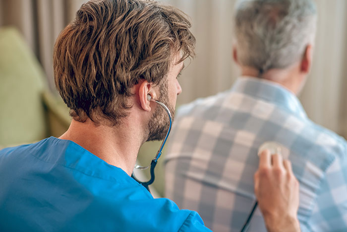 Young male doctor in blue scrubs using stethoscope to examine elderly man, illustrating health in adulthood for ultra-popular guys.