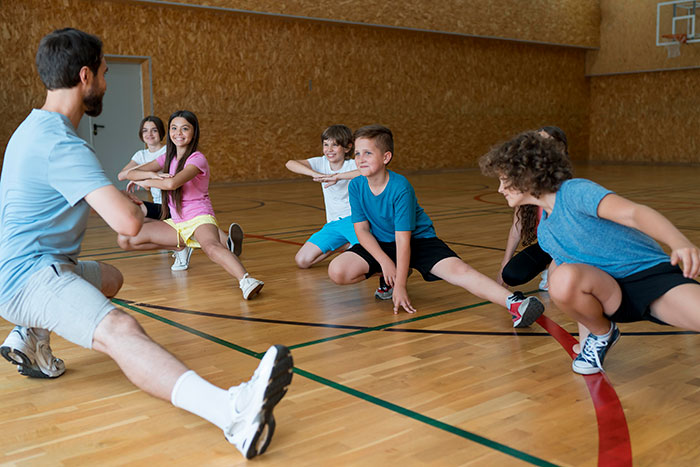 Group of kids stretching with male coach in gym, illustrating active lifestyles and social development of popular guys.