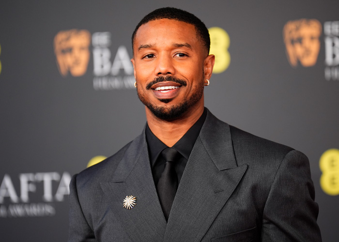 Michael B. Jordan smiling at an event, dressed in a dark suit, fans stunned by his transformation for Sinners Twins role