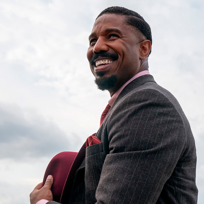 Michael B. Jordan smiling outdoors wearing a suit, showing the transformation he did to his face for Sinners Twins role.