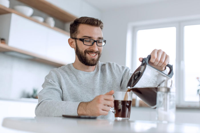 Man pouring dark roast coffee into a glass mug in a bright kitchen, smiling while enjoying his daily coffee ritual.