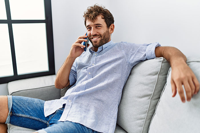 Smiling man sitting on couch making a phone call, representing husband lied during video call before divorce.