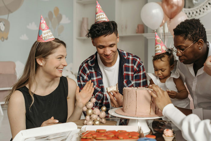 Family celebrating birthday with cake and party hats as lady throws big party for hubs despite his feelings.