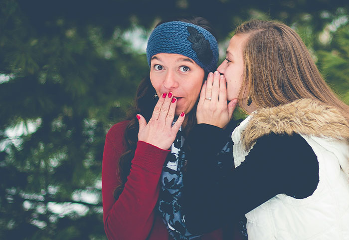 Two women outdoors, one whispering a secret to the other, illustrating unhinged workplace incidents and surprising confessions.