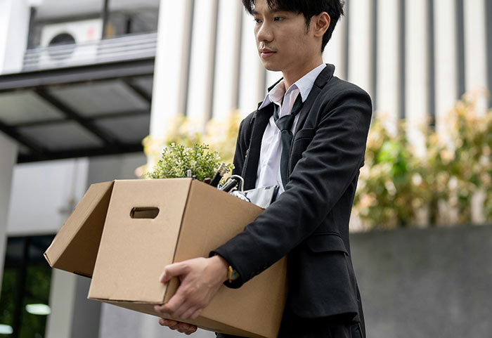 Young professional in a suit carrying a box of personal items, illustrating unhinged workplace incidents and job challenges.
