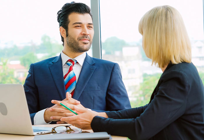 Two professionals in a business meeting discussing workplace incidents and office challenges near a window with city views.